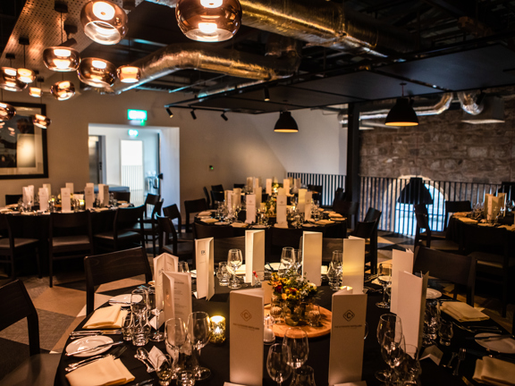 A view of a private dinner mezzanine space at the Clydside Distillery, filled with round tables with black table cloths. They are decorated with floral centrepieces, candles, silverware and menus. Wooden chairs with soft seats are arranged around them. What floor is visible is patterned with a checkerboard effect, but in the low light of the room it is not possible to discern the texture. A black railing runs the width of the mezzanine, a bare stone wall with an arched window is visible beyond.