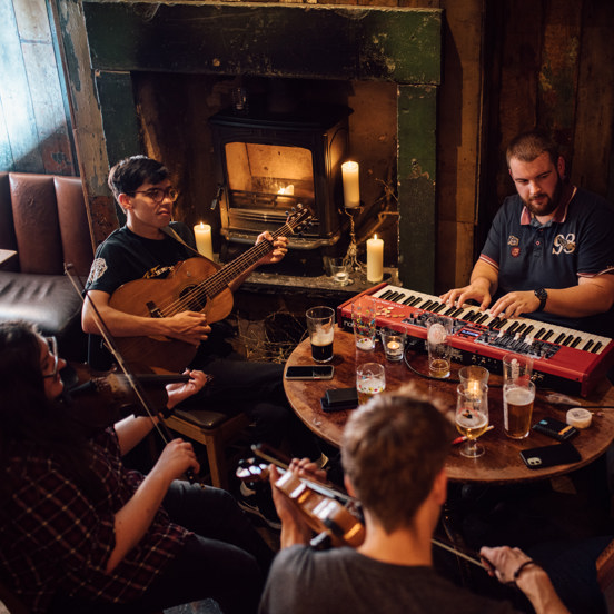 group of 5 musicians  around a table in a restaurant playing instruments, with several pints on the table and fire in the background