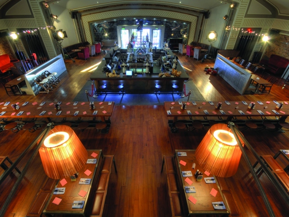 An interior view of the Grosvenor cafe shows a large room with a wooden floor and a high, arched ceiling. The photo is taken from above, looking down on multiple long, rectangular wooden tables with stool and bench seats. At the back of the space is a large, square bar with shelves in the centre and on beyond it on the far wall is a window. The room is otherwise lit with lamps, and fairy lights. Sofas and seating can be seen around the edge of the space.