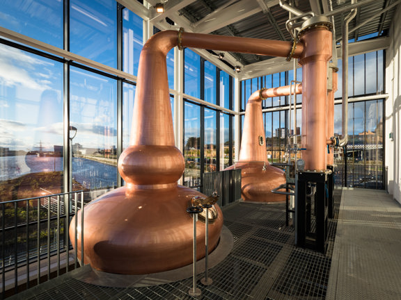 A view from inside the Clydeside Distillery. A room with glass walls, it is taken in daylight from a first-storey mezzanine. The space is dominated by 2 enormous, copper whisky stills - an important instrument in the distilling of whisky. They have bulbous forms and highly polished surfaces, copper and silver pipes lead off of them. A black railing runs the length of the mezzanine, the floor is a mix of metal grates and tread plate. Out of the windows views of the River Clyde and blue skies are visible. 