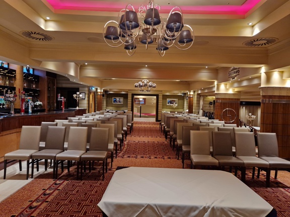 An interior view from Ingliston Country Club shows a room decorated with an ornately patterned orange and maroon carpet. There is a long polished-wood bar along the left-hand wall. The ceiling is ornately plastered, lit pink with LED lights and decorated with a spiralling silver ceiling light, small black fabric lampshades cover the lightbulbs. The floor is filled with about 10 rows of grey, leather-effect high backed chairs. At the back of the room, centred with the chairs aisle is a large open doorway.