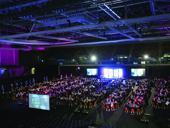 Interior image of the main hall of the Emirates Arena. The picture shows a large dark hall, lit by projections and coloured lighting effects. The space is filled with dozens of round tables with black table cloths and red velvet chairs surrounding them. There are 3 large projection screens visible around the edges of the seating. Empty spectator seating can be seen on the upper level of the arena. The ceiling is obscured by technical light and camera scaffolds. 