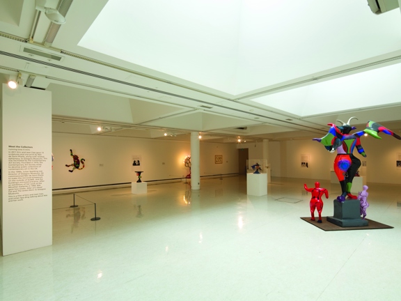 An interior view from GoMA shows a large, bright, white gallery space. There is evidently sky lights in the roof as well as spot lights in the ceiling. The floor is highly polished and dotted with plinths and brightly coloured sculptures. A pillar can be seen with interpretive text on it and further artworks can be seen on the walls. A doorway leads out of the room in the back corner of the space.