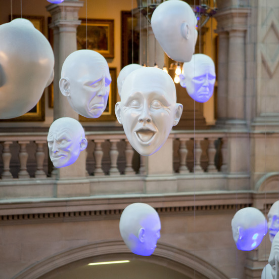 art exhibit consisting of white acrylic heads with expressions of various emotions, hanging from the ceiling in the Kelvingrove Art Gallery and Museum