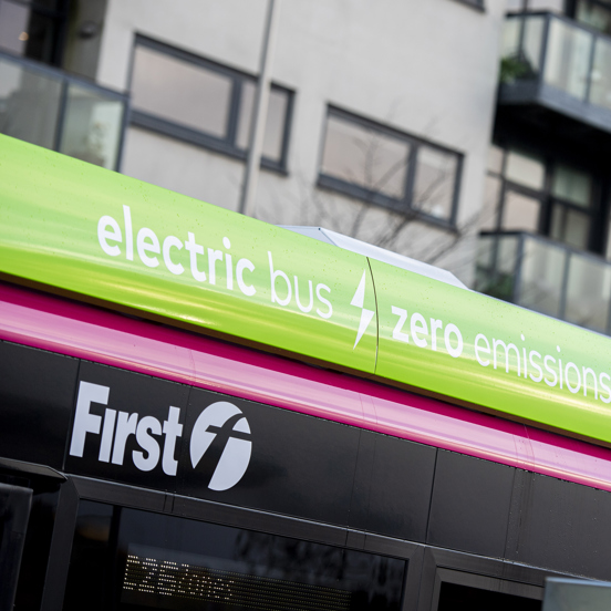 Section of a bus, with First Bus logo, and electric bus signage, with a bus stop sign in the foreground
