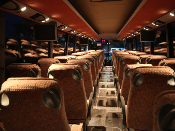 An image taken looking down the length of a luxury coach from the back seat. The seats on either side of the aisle are upholstered in orange fabric and have armrests. The floor is carpeted and the ceiling has orange panels with small spotlights above each seat. 4 small screens can also be seen attached to the ceiling. Outside is dark so the coach's interior is reflected in the large windows.