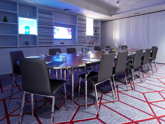 An interior photo of a Malmaison meeting space. A series of chrome and wood-effect tables have been positioned side by side to create a long board room table. Sleek chrome and leather chairs surround it. The room is darkened, predominantly lit by a large TV screen and a light box on the far wall. The TV is surrounded by a shelved unit, small items -vases and a globe- are sporadically placed on the shelves. The shelves, walls and ceilings are white, while the carpet is pale grey with a geometric red pattern.