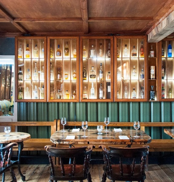 An interior view of a dining room at The Finnieston restaurant. A bright room with refinished wooden floorboards and walls decorated with reclaimed wood panelling and green corrugated metal. Light varnished pigeonholes line the far and left walls, they are lit and glass fronted – displaying bottles of whisky. A long varnished wooden bench, that lines the base of both walls, and dark wood, round-back chairs are arranged around wooden tables, with cast iron legs, set for 2 – 4 people.
