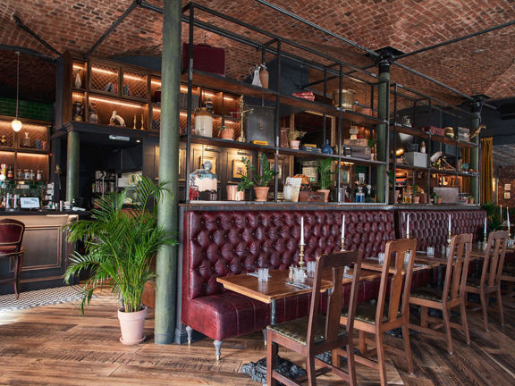 An interior view of the Duke's Umbrella bar and dining room. The space is predominantly floored with varnished wood, with checked tile patches in front of the dark panelled bar. A long, high-backed red bench forms a barrier between the bar and dining space. Small wooden bistro tables with iron bases line its length, with a wooden dining chair at each table. Shelves above the bench and behind the bar are decorated with plants and eclectic ephemera. The ceiling is exposed brick, supported by 3 green pillars.