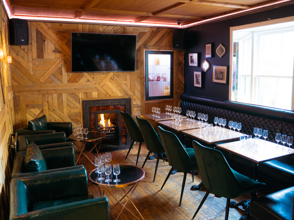 An interior view of a space at The Finnieston restaurant, arranged for a private drinks event. A small room with reclaimed wood panelling on the left and far walls, a large TV is mounted to the wall above a small, lit fireplace. The right hand wall is painted dark grey; a large, bright window and small, eclectic frames decorate it. Leather armchairs, velvet dining chairs and a long leather studded bench make up the seating. Low copper coffee tables and wooden dining tables are arranged with wine glasses.