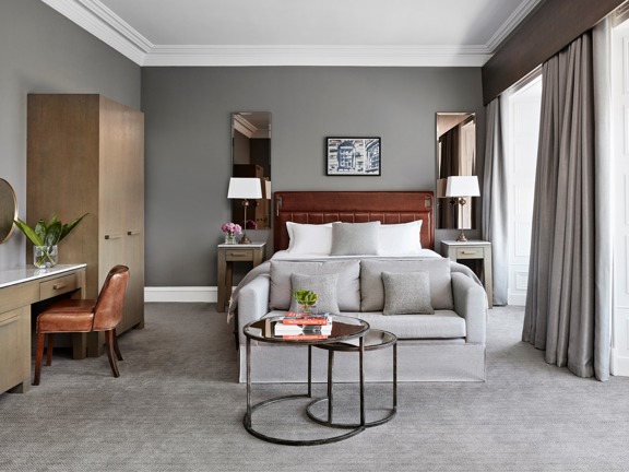 An interior shot of a Blythswood Square hotel room, a room with soft pale-grey carpet, grey walls and white plaster work, 2 windows surrounded by white wooden panelling are evident on the left wall. A large bed with a brown leather headboard made with crisp white sheets is against the far wall. a pale grey loveseat is at its foot. 2 small, round glass nesting coffee tables, with black metal bases, sit in the foreground. Against the left wall are a modern, ashen-wood wardrobe and desk & a leather chair.