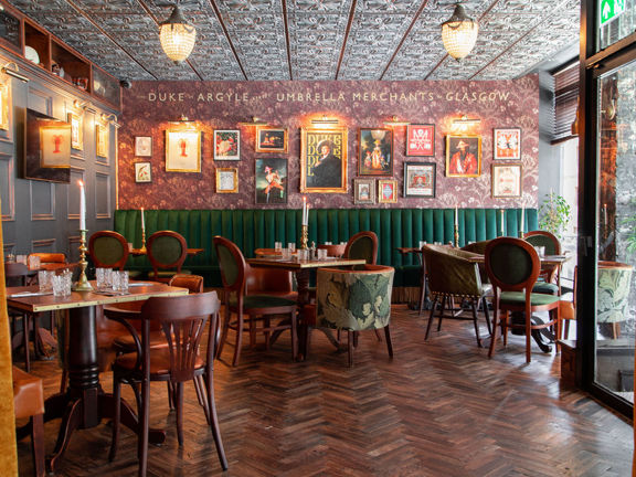 The richly decorated dining room of the Duke's Umbrella. The restaurant is darkly decorated with dark-wood parquet flooring and panelling. Deep red botanical wall paper decorates the far wall, with gilt framed prints and brass spotlights curated on it. A high-backed deep-green upholstered bench lines the far wall. The room is otherwise furnished with dark wood tables and eclectic chairs. Plate glass windows allow daylight to stream in, while cut-glass light features are mounted to the carved-wood ceiling.