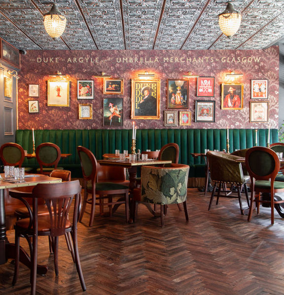 The richly decorated dining room of the Duke's Umbrella. The restaurant is darkly decorated with dark-wood parquet flooring and panelling. Deep red botanical wall paper decorates the far wall, with gilt framed prints and brass spotlights curated on it. A high-backed deep-green upholstered bench lines the far wall. The room is otherwise furnished with dark wood tables and eclectic chairs. Plate glass windows allow daylight to stream in, while cut-glass light features are mounted to the carved-wood ceiling.