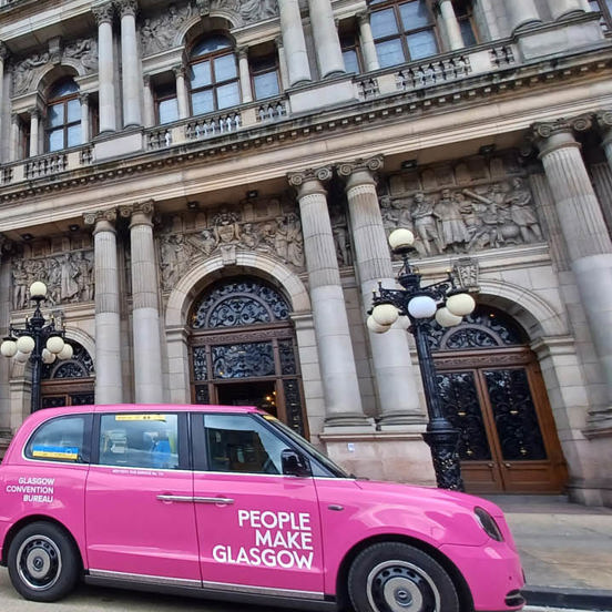 3 pink taxis with People Make Glasgow branding lined up in front of the Victorian-style Glasgow City chambers