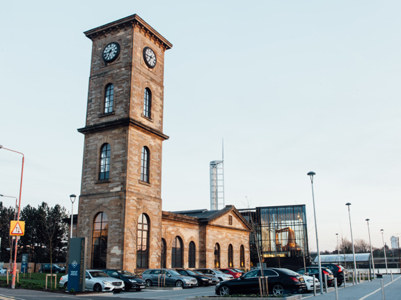 An external view from the carpark of the Clydeside Distillery. A tarmacked carpark wraps around an old sandstone building. It has a large square clock tower on the nearest end with arched windows up its sides. The rest of the old building is single storied with large arched windows flanking its sides. A modern glass extension can be seen over 2 floors at the back, right of the building. The Glasgow Science Centre tower can be seen behind the roofline of the Clydeside Distillery against a pale evening sky.