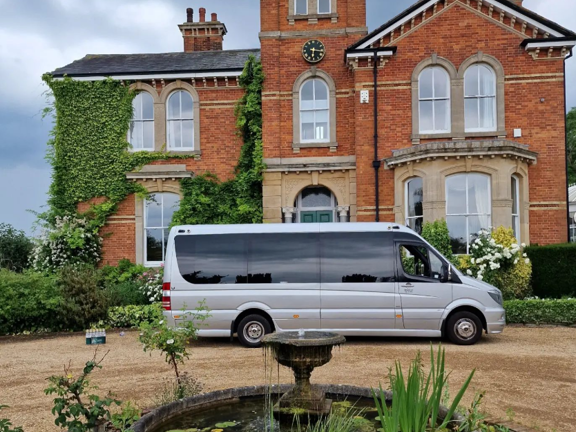 A large silver people-carrier with dark windows can be seen parked on the circular gravel driveway of a large brick house. Ivy climbs the right edge of the house, shrubs line the drive and an ornamental fountain with water lilies and irises is in the centre. The sky is cloudy and grey. 