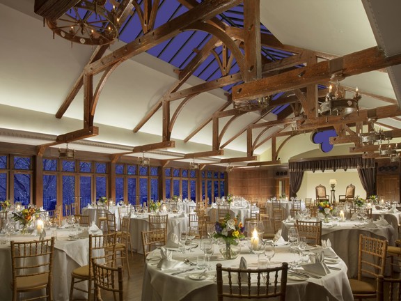 An interior view of Crossbasket's event space shows a long room with a vaulted ceiling. Wooden beams are visible the length of the ceiling, in the roof there is a skylight and the longest wall is also windowed. At the far end of the room a curtained stage is visible. Throughout the room large round tables and chairs are decorated with flowers, candles, wineglasses and silverware.
