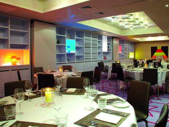 An interior photo of a Malmaison private dining space.  Sleek chrome and leather chairs surround round tables, with white tablecloths and candles, are laid for a dinner. The room is darkened, predominantly lit by a modern ceiling light and coloured lights on the far wall. The lights are surrounded by a shelved unit, small items -vases and a globe- are sporadically placed on the shelves. The shelves are pale grey & the walls and ceilings are white. The carpet is pale grey with a geometric red pattern.