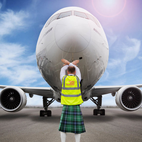 Back view of a man in kilt and yellow hi-vis jacket with People Make Glasgow branding in front of a large plane