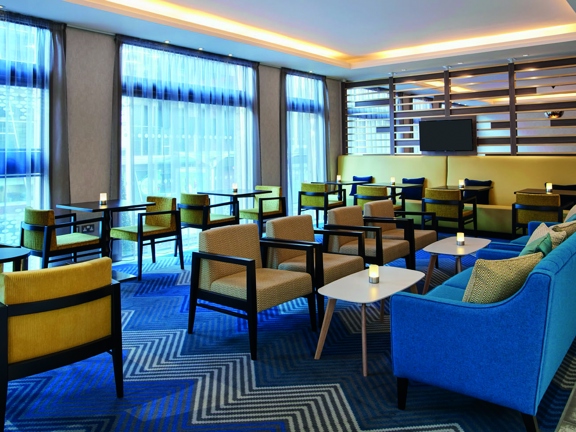 An interior view of the Hampton by Hilton lobby, a bright room with large floor to ceiling windows covered with net curtains is decorated with a bold zig zag rug and wooden floors are visible. Small dark tables with plush yellow chairs are arranged in the space, each candle has a small lantern. 2 teal sofas and accompanied by small teak coffee tables on the right while a plush yellow bench and a slatted wooden wall create a divider to the rest of the room. A TV screen can be seen on the slatted wall.
