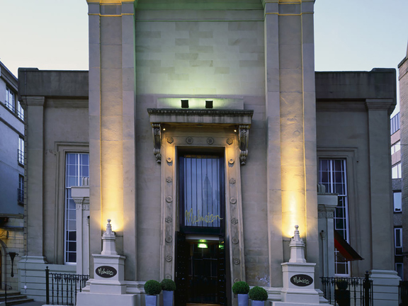 An exterior image of the Malmaison Glasgow shows a sandstone building with a strikingly, tall Egyptian-influenced façade. An enormous, carved archway houses the entrance, a black double doorway with a large window above it. Gold lettering above the door in the signature branding reads, "Malmaison." The façade is uplit with white and green lights, exaggerating its height and showcasing the carved pillars and a Greek inscription at the very top. Black railings and potted shrubs decorate the steps.