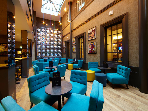 An interior view of the Malmaison Bar, shows a narrow, double-height room with a sky-light roof. A metallic art work of discs takes up most of the far wall. Low, velvet chairs and footstools in turquoise, teal and yellow surround small dark, bar tables. A lower ceilinged seating area can be seen through 2 mirrored pillars to the left. Framed prints and dark-wood internal windows and a picture rail adorn the right wall.