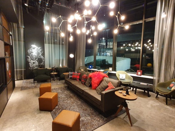 An interior view of Moxy's lounge area shows an open space with floor to ceiling windows on the right-hand exterior wall. The floor is polished concrete with grey textured rugs in parts. A eclectic variety of armchairs, sofas and footstools are spaced out, with coffee tables. The walls, where they are not windowed, ae painted dark grey. There is a modern, branching ceiling light, brightly lighting the room; the far wall has a mural of musical instruments. Industrial-style shelving can be seen on the left.