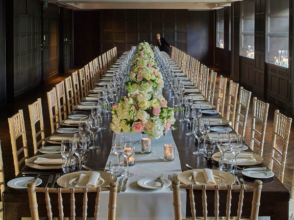 An interior from the Blythswood hotel of a room arranged for a private dinner event shows an incredibly long and ornately decorated table. 2 dozen spindle-backed light-wood chairs surround a table adorned with bouquets of pink and white roses, candles, silverware and glasses. 4 windows are evident along the right hand wall, brightening the dark wooden panelling present on all of the visible walls. Arrangements of candles sit in each of the window's alcoves.