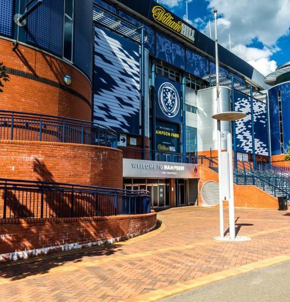 An exterior view of Hampden Stadiums main entrance. A large brick and blue metal clad building, with round towers at the two visible corners. The facade is decorated with sponsors, the Scottish FC badge and lettering in white or yellow reading," Hampden Park."  The area outside of the main entrance is paved with brick and curves down to 3 or more doors. Adjacent steps lead up to a further entrance, furthermore, ramps curve out of sight around the towers' bases. Trees and modern streetlights line the path.