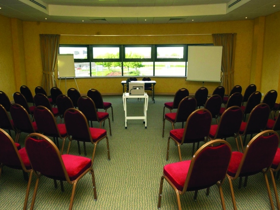 An interior shot taken from the back of a meeting room at the Campanile Hotel; with striped carpet and cream walls. At the far end of the room a row of dark-framed windows gives a view of a landscaped bank of trees and the SEC campus beyond. There is a projection screen on a stand on the right, a table with 2 chairs in the centre and a flip chart on the left. 4 rows of red velvet chairs take up most of the room. A projector is on a wheeled trolley in the aisle. Spotlights can be seen in the ceiling.