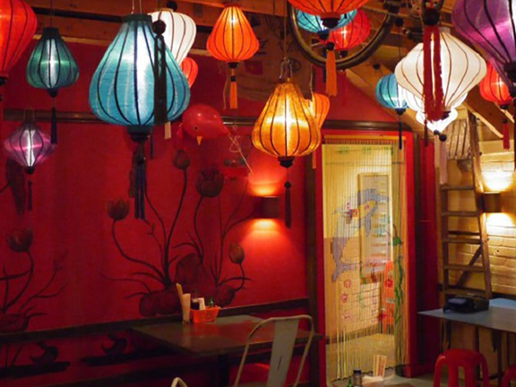 An interior image of Hanoi Bike Shop shows a small, low ceilinged dining room, the walls are painted a rich red and decorated with hand-painted floral motifs and lettering. Lantern light fixtures in an array of colours hang from the ceiling. Small tables and an eclectic mix of stools and chairs can be seen around the edges of the room. A beaded curtain covers an open, brightly lit doorway in the back corner of the room.
