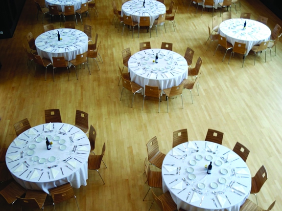 An internal view of the Maryhill Burgh Hall is taken looking down at a large hall. Large round tables with white table cloths are surround by a dozen wooden wire-legged chairs. The tables are laid for dinner with silverware, crockery and bottles of wine visible. The floor is light-wooden floorboards and at the far end the edge of a small, temporary wooden stage is visible. The lower half of a dark-wood panelled wall can just be seen. The space is brightly lit, suggesting the room has large windows.