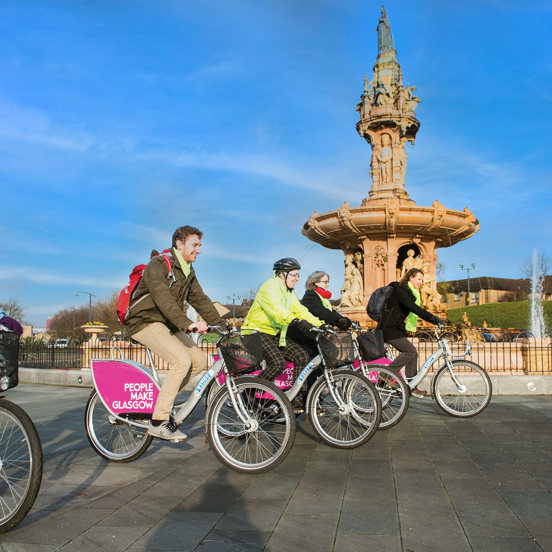 Five cyclists on People Make Glasgow branded bikes in front of the large terracotta Doulton Fountain