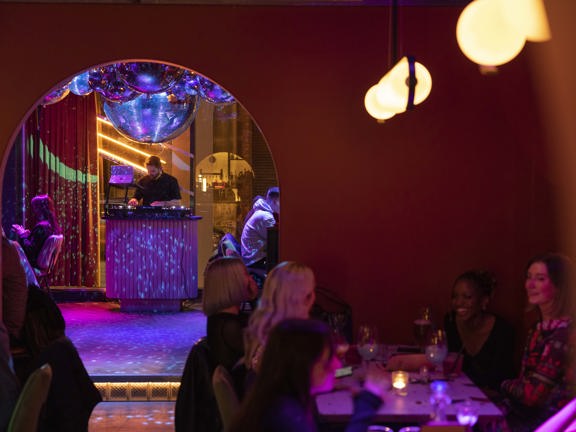 An interior view of August House, two rooms are joined by a wide arched doorway, with a small stop up. Through the archway a DJ can be seen at a small semi-circular podium. Lights and mirrored disco balls project specks of blue light across the floor and podium. Customers sit at tables with drinks in front of them. The closest room has low lighting form over head spherical light fittings and small tea light candles.