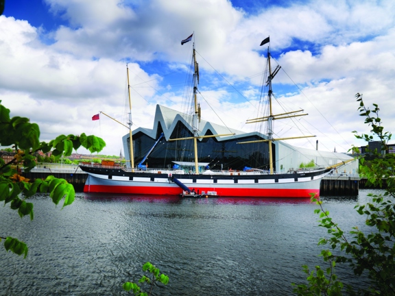 A view of the Tall Ship taken from the south bank of the River Clyde, shows a large moored ship painted with horizontal stripes of black, white, grey and red. The ship has 3 large, yellow masts with rigging and flags flying. Behind the ship is the tall, silver, zig-zag roofline of the Riverside Museum. Trees branches flank the photograph on either side of the frame and a blue, cloudy sky and the tops of a few buildings can be seen in the background.