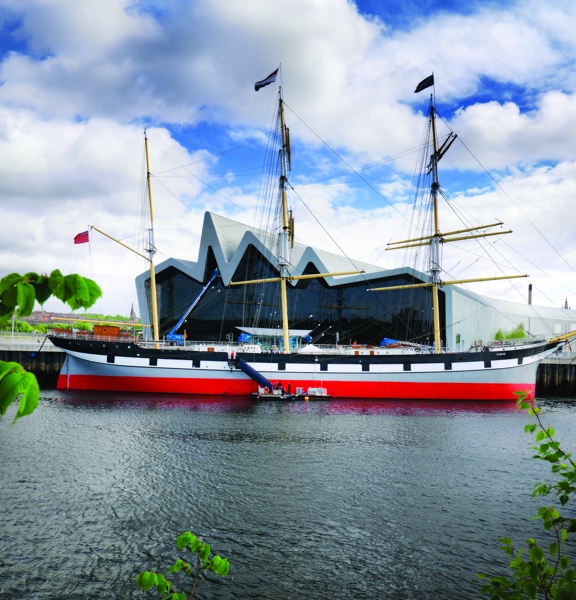 A view of the Tall Ship taken from the south bank of the River Clyde, shows a large moored ship painted with horizontal stripes of black, white, grey and red. The ship has 3 large, yellow masts with rigging and flags flying. Behind the ship is the tall, silver, zig-zag roofline of the Riverside Museum. Trees branches flank the photograph on either side of the frame and a blue, cloudy sky and the tops of a few buildings can be seen in the background.