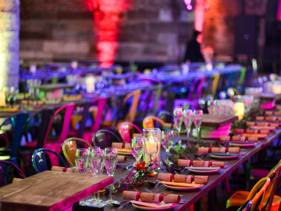A detail of a function table set-up at Platform, a long table is decorated with cut flowers, champagne flutes, candles and Christmas crackers. Yellow and black glossy metal chairs line both sides of the table. The space is dark and lit by coloured red and pink lights which shine on the bare stone walls. In the background a second long, decorated table is pictured out of focus.