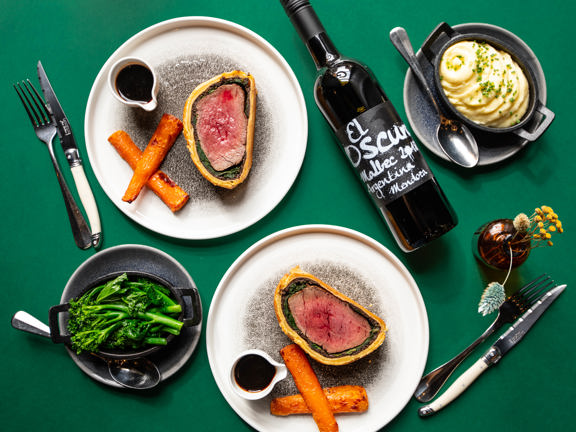 A stylish "flat-lay" photograph of a Glaschu dining table. A table laid with a dark green cloth is decorated with 2 minimal plates of beef wellington, roasted carrots and small jugs of gravy. Rustic sides plates hold creamed potatoes and steamed green vegetables. A bottle of red wine, silverware and a small brown glass vase of dried flowers complete the arrangement.