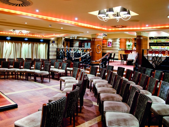 An interior view from the Ingliston Country Club, shows a space over 2 levels. The lower level is decorated with a dark-wood parquet floor, plush wooden and grey velvet chairs are arranged in semi-circular rows facing a small stage. The ceiling has plastering details and is lit orange with LED lighting The few stairs and the upper level are carpeted, ornate railings and pillars separate the levels. A long lit bar is visible in the background.