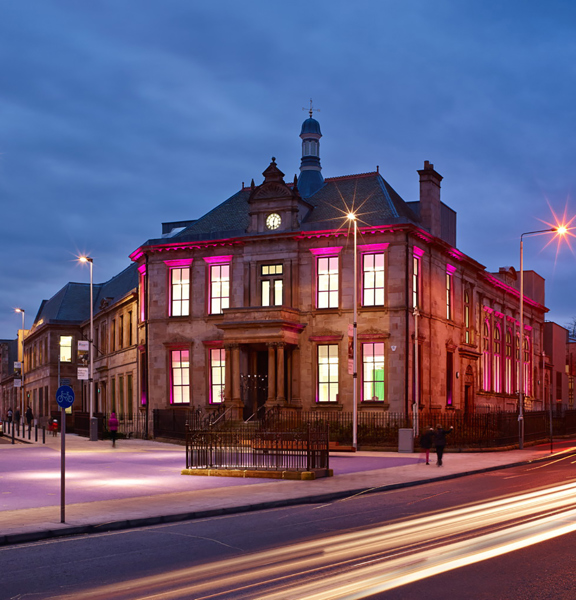 An exterior view of the Maryhill Burgh Hall shows a Victorian, 2-storey municipal sandstone building on a street corner, with a grand pillared entrance. The image is taken at dusk with a long exposure so streaks of light fill the road. The large windows are lit and the building is up-lit with pink lights. Streetlights, parked cars, tenements and a railway bridge can be seen.