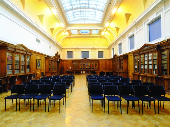 An interior view of a large room in the Mitchell Complex. Textile and metal stacking chairs are arranged into rows on a polished, parquet floor. The room is bright with a large skylight taking up most of the arched, decoratively plastered and painted ceiling. The walls are wood panelled to half their height, the upper portion has windows, plaster work and alcoves decorating them. Ornate, glass-fronted shelves and display cabinets line the walls and a guilt framed painting is also visible on the left wall.