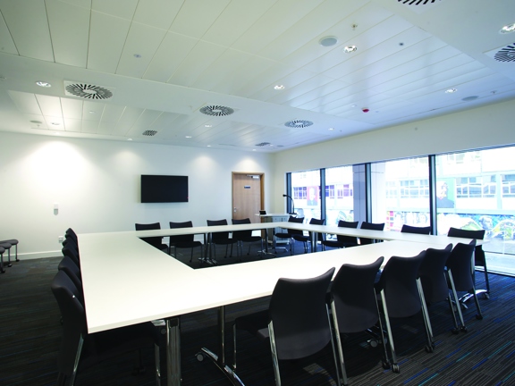 The interior of a private meeting space at the TIC, the room is painted white with a dark, striped carpet. The exterior wall to the left has floor to ceiling windows and looks out onto George Street and the Strathclyde University Mural. White tables with metal bases are arranged into a large square. Dark chairs with metal legs line the outside edge. A large television screen is on the far wall to the left of a doorway. The ceiling is white and panelled, with frequent spotlight and vents.