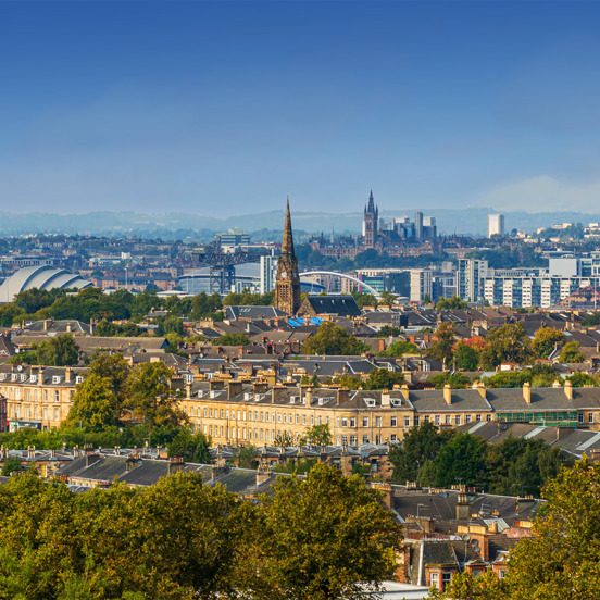 Sunny cityscape with the Gothic revival-style spire of the University of Glasgow's main building, surrounded by greenery and buildings of the West End, including the modern buildings of the SEC Armadillo and the OVO Hydro arena