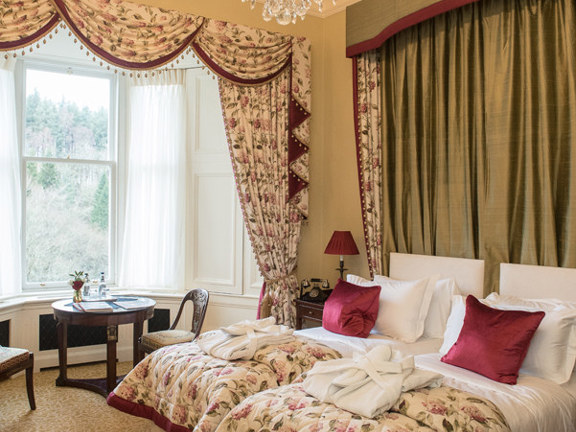 An interior shot of a twin bedroom at Crossbasket Castle hotel. The room has 2 single beds, positioned closely together to the left of the image. The beds and room are decorated opulently with floral throws, curtains and pelmets in cream and magenta. On the end of each bed is a white, plump, folded dressing gown. The bay window has white net curtains and a small round table and 2 wooden chairs sit in the bay. A lit chandelier hangs above the beds and illuminates the plaster ceiling-rose.