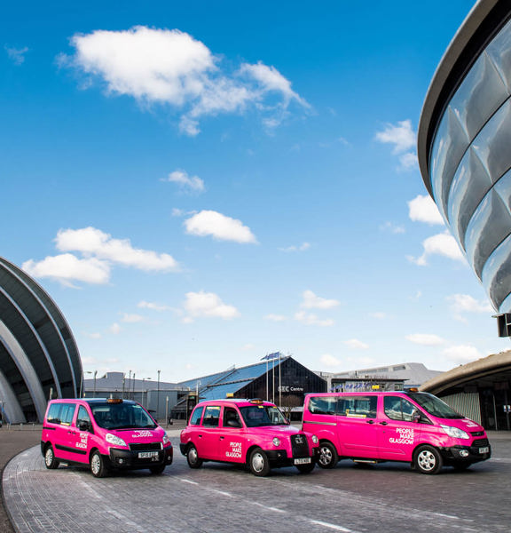 3 pink taxis with People Make Glasgow branding lined up in front of the modern structures of the Scottish Event Campus