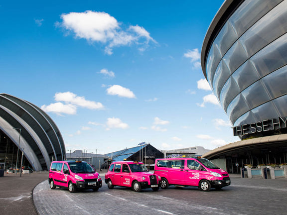 3 pink taxis with People Make Glasgow branding lined up in front of the modern structures of the Scottish Event Campus