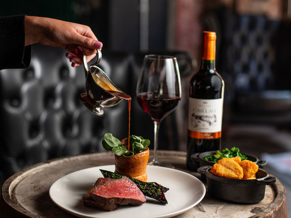 A close up image of a Duke's Umbrella roast dinner placed atop a reclaimed wooden barrel. A white, feminine hand and fore-arm can be seen pouring gravy from a small silver gravyboat onto a clean plate of roasted vegetables, sliced beef and Yorkshire pudding. Rustic metal bowls of roast potatoes and greens, a bottle of red wine and a large wine glass can also be seen on the wooden top. Plush, black leather seating is visible in the background.