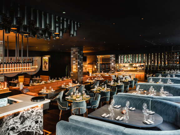 An interior view looking down on Gaucho's expansive dining room. The space is darkly painted, with furnishings and accents in gold, grey, bronze and marble. A dramatic, monochrome marble bar is visible to the left, while seating to the right and ahead is varied, a mix of booths, benches and chairs around grey or gold topped tables of different sizes. The tables are set with tall wine glasses, silverware and cloth napkins. Lighting away from the windows is low and golden.