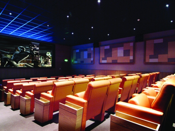 An interior view from the aisle of the Grosvenor Cinema shows a small cinema screen and seating. Several spotlights in the ceiling light 8 rows of orange, leather fold down chairs. An animated image can be seen on the wide screen and geometric artworks in orange are on the far wall. In the far corner a fire exit sign is visible.