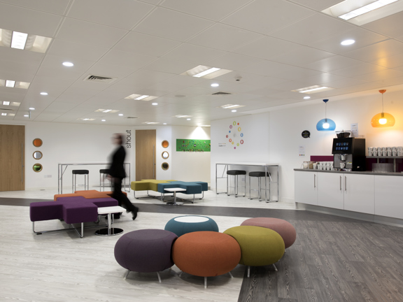 An photo of The Studio's break hall shows a large, bright interior space with white walls and wood effect flooring. Small islands of modern, brightly coloured seating in round and cross-shaped fabric pouffs are dotted throughout the space. The right hand wall has a long catering bar, with a coffee machine, mugs and tap. Glass blue and yellow ceiling lights hang above it. 2 high tables with stools are seen against the remaining wall space. A corridor and 2 doors can be seen off of the far wall of the space.